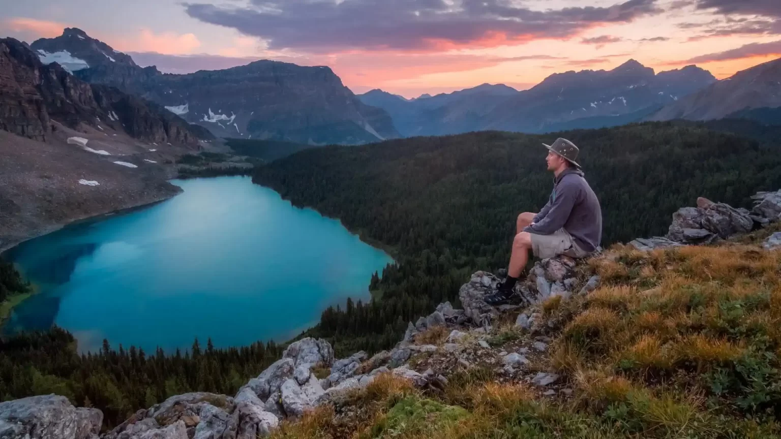 A person sitting on a mountain overlooking a lake, forest, other mountains and a sunset