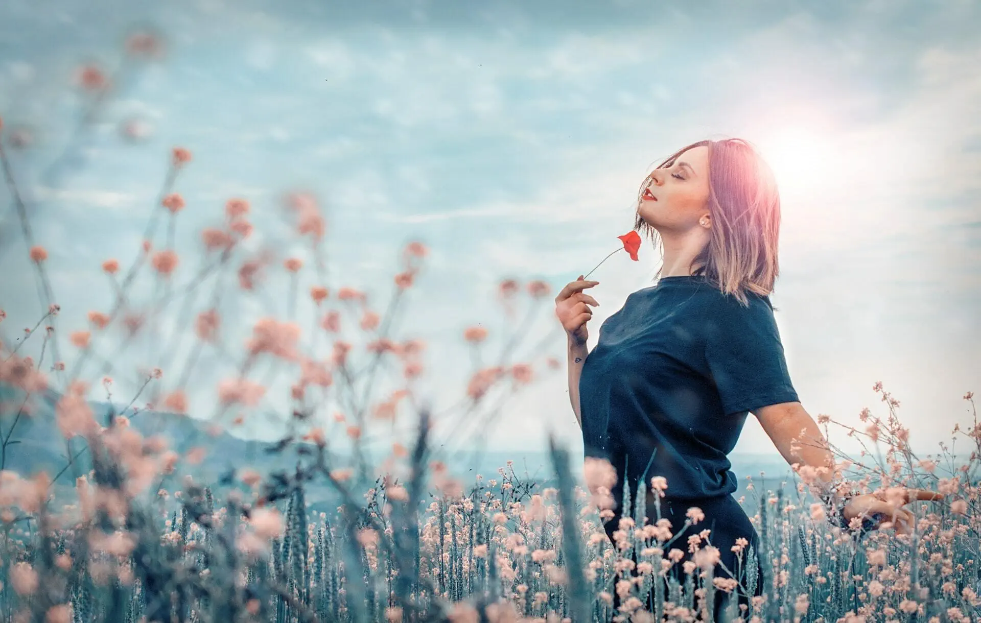 A woman holding a flower in a field, with her eyes closed and head up to the sky